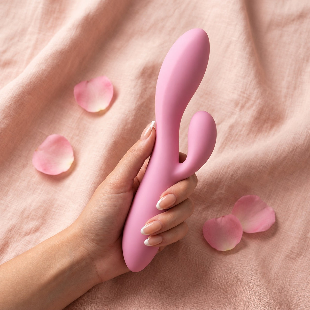 Hand holding the pink LoveLab Symphony rabbit vibrator diagonally on pink linen with rose petals